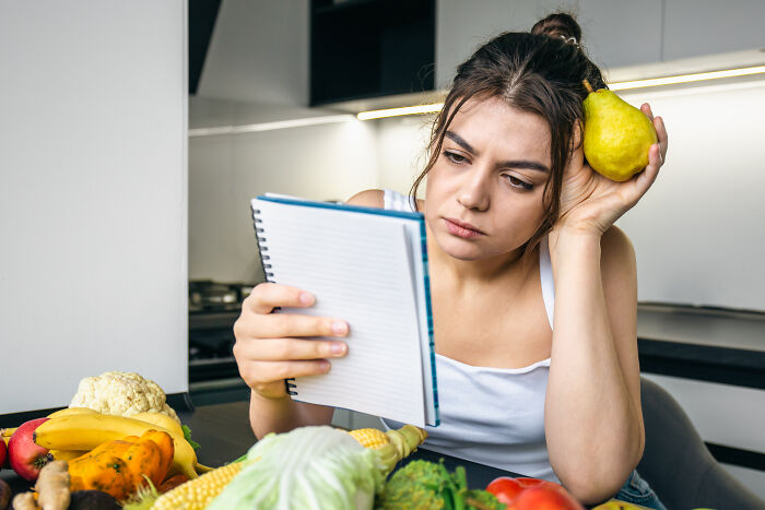 A woman looks confused while holding a pear and a notebook. She is surrounded by healthy foods, like vegetables and fruits.