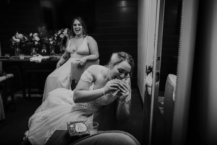 A bride laughs while eating a burger in this Funny Wedding Photos moment, with another woman laughing in the background.
