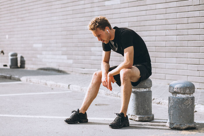 A tired man in athletic wear sitting on a stone post, reflecting on healthy habits, exercise, and the need for rest.