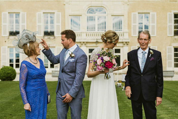 A wedding party posing outside a grand building, with a groom and bride comically poking family members, capturing street photography.