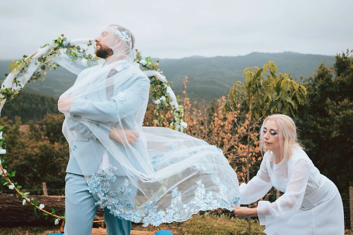 A man has a veil over his head while a woman holds it, a funny wedding photo outdoors.