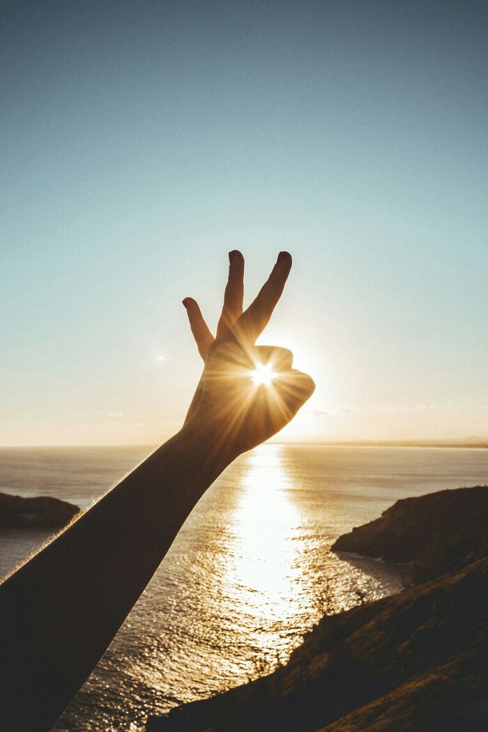 A hand forming a peace sign against a bright sunset over the ocean, symbolizing healthy habits and a positive lifestyle.