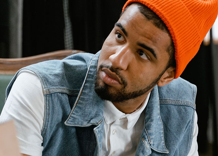 A man in an orange beanie and denim vest looking away, possibly reflecting on his worst first dates.