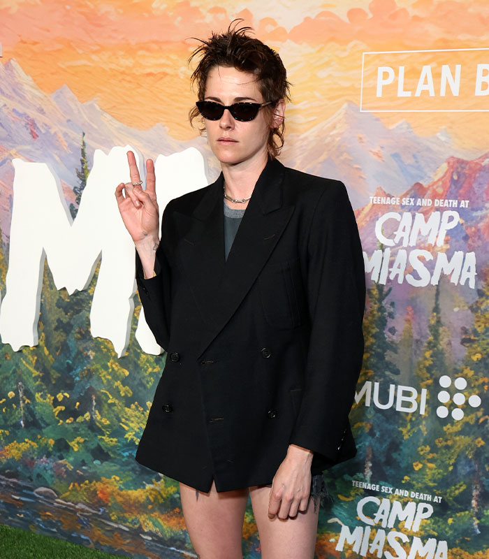 Kristen Stewart at an event, wearing a black blazer and sunglasses, making a peace sign, for her unsettling appearance.