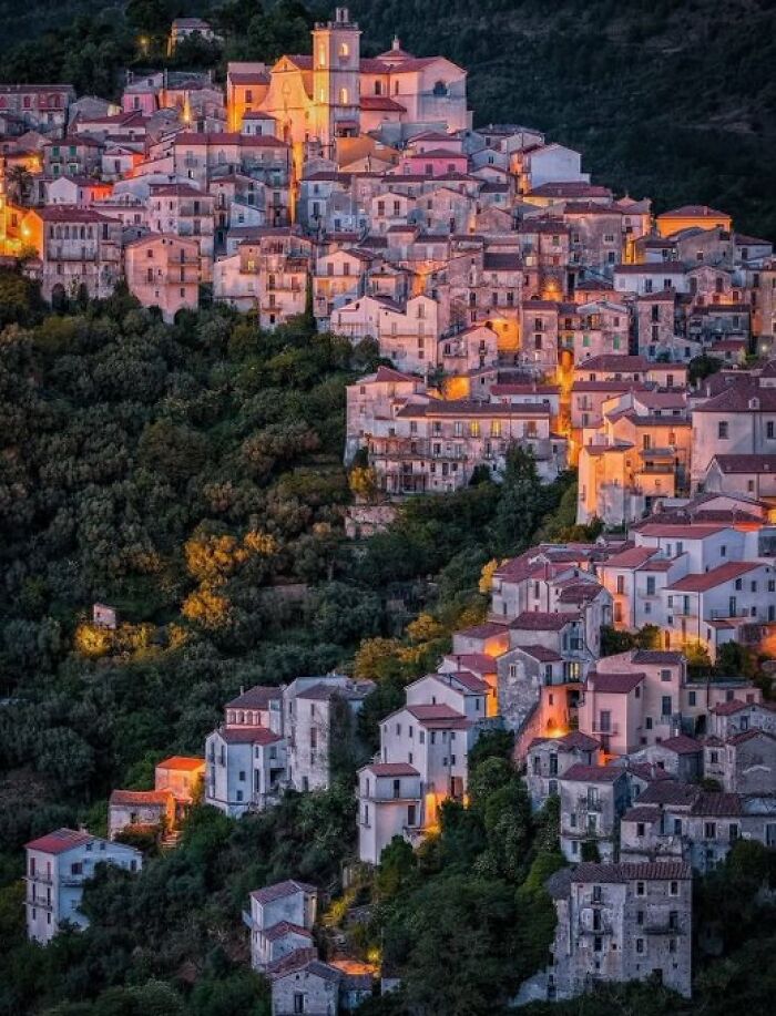 A charming hillside town lit up at dusk, with numerous houses nestled among trees. Buildings with so much character.