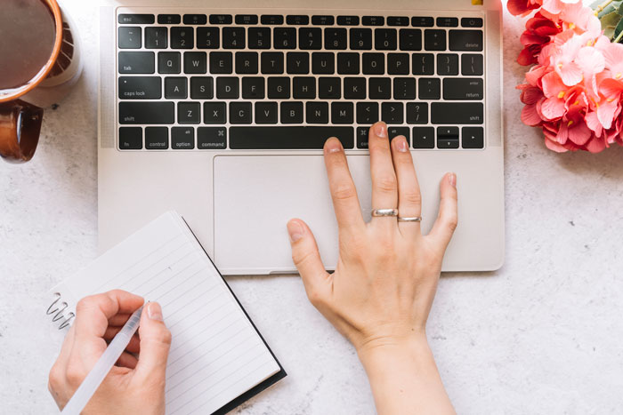 Hands typing on a laptop and writing in a notebook, researching things that happened at weddings.