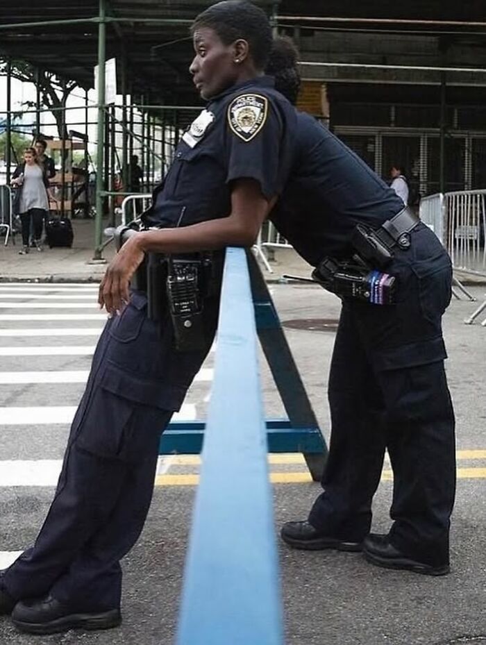 A photographer captures a funny moment of two police officers leaning on a blue barrier, one with her head on the other's shoulder.