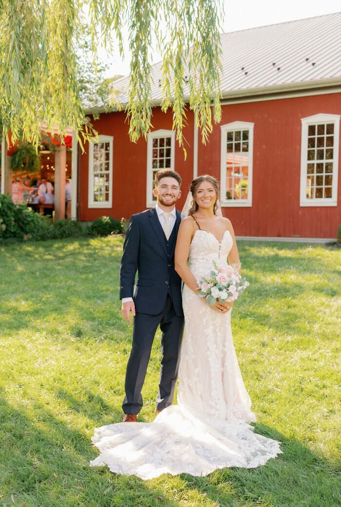 A couple smiles in front of a red barn, a funny wedding photo with a beautiful backdrop.