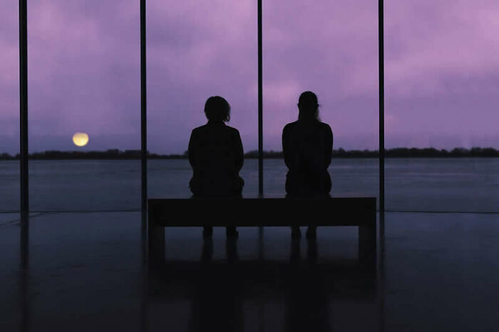Two figures sit on a bench facing a window with a dramatic purple sky and moon, a cinematic portrait.