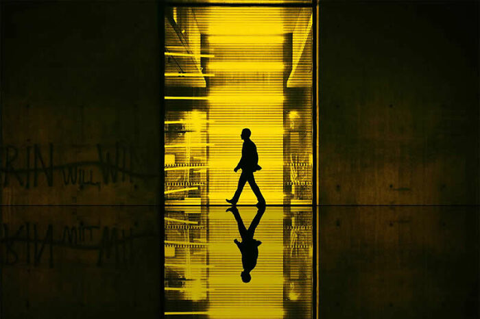 A silhouette of a person walks across a bright yellow lit pathway, reflecting below, in a dramatic example of cinematic portraits.