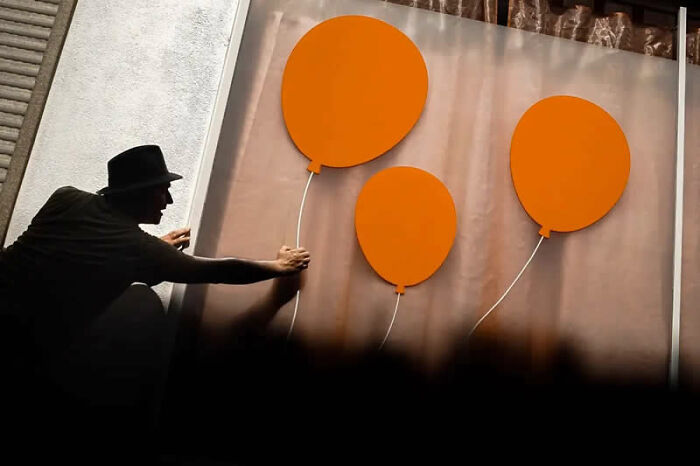 Best street photos from the Macadam Awards show a man in a hat reaching for orange balloon cutouts on a wall.