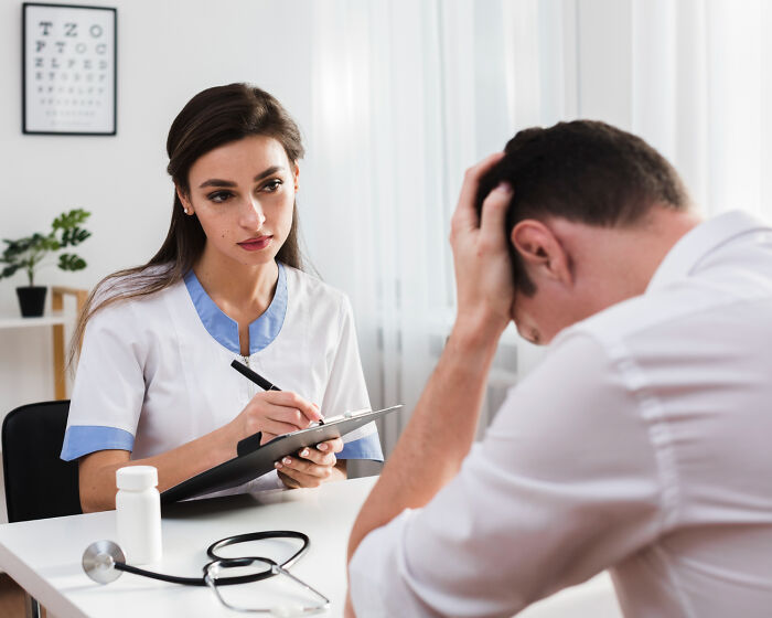 A female doctor taking notes while a male patient holds his head, discussing difficult diagnoses.