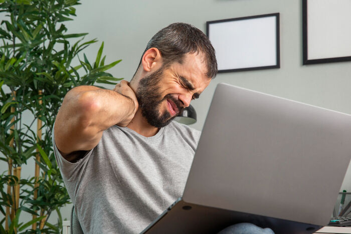 A man with a beard grimacing while rubbing his neck, working on a laptop. Doctors refuse to do some things.
