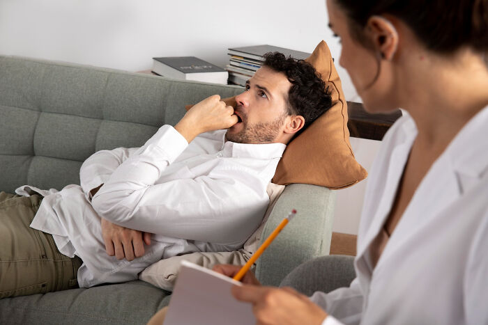 A man lies on a couch, looking distressed, while a therapist takes notes, exploring challenges in therapy.