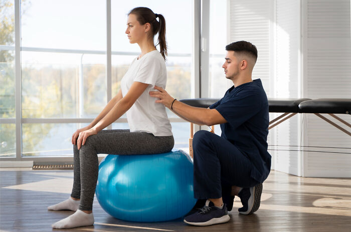 A physical therapist assists a female patient on a blue exercise ball, focusing on posture and core strength in the medical field.