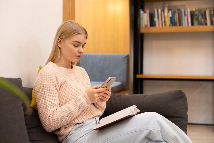 A young woman in a pink sweater sitting on a couch, holding her phone. She walked into therapy and walked out without her therapist.