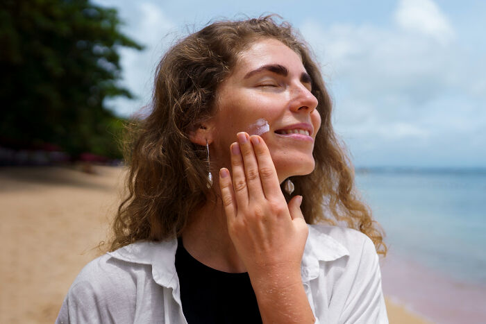 A smiling woman applies sunscreen to her face at the beach, a vital self-care step doctors refuse to skip.