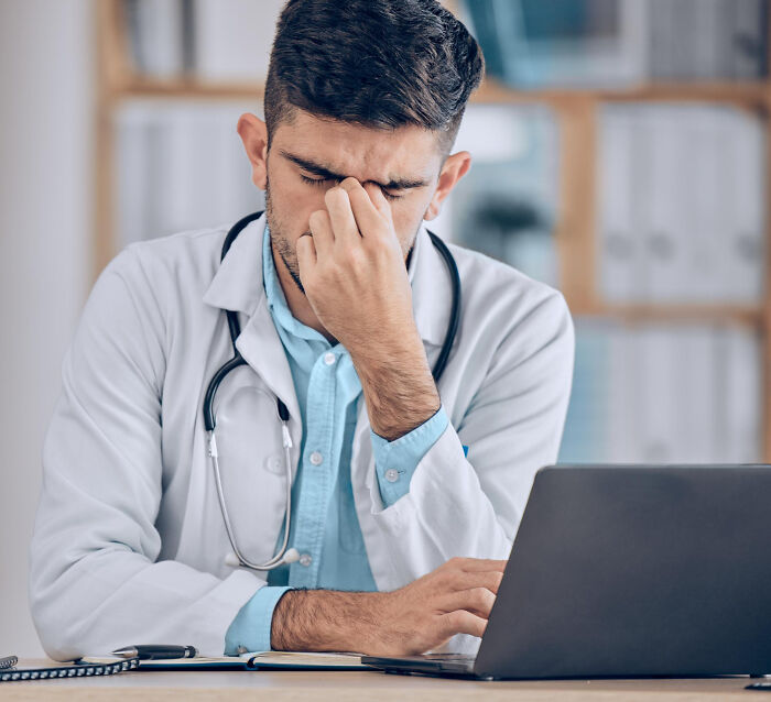 A medical field worker, dressed in a white lab coat and stethoscope, looks stressed while working on a laptop.