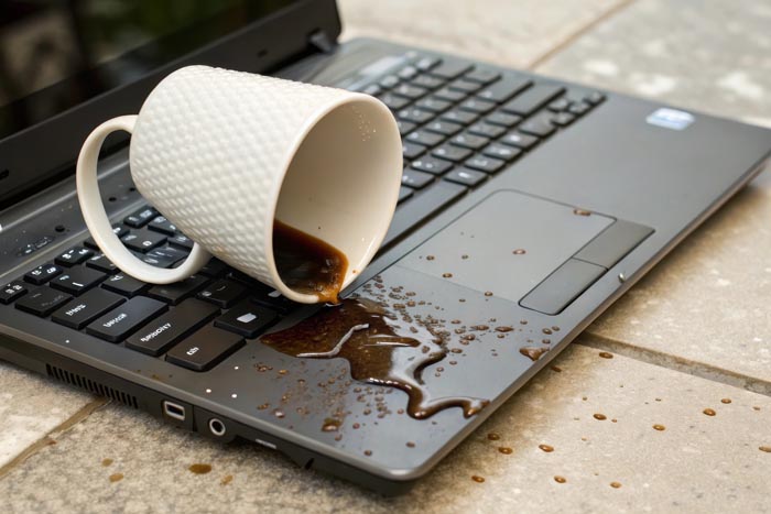 A white mug spilling coffee onto a laptop keyboard, representing dumb work rules.