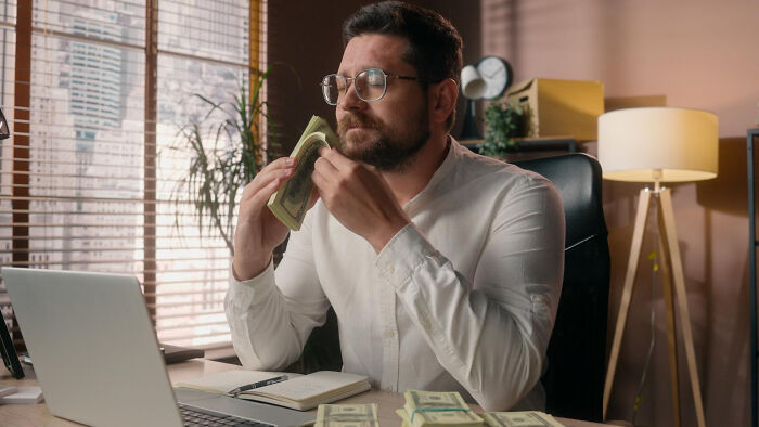 A man in a white shirt with glasses smelling a stack of money at his desk, highlighting dumb reasons couples divorce.