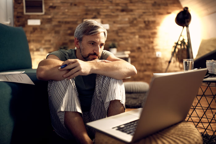 A millennial man with a beard sits on the floor, looking at his laptop, considering new crisis hobbies. A millennial man with a beard sits on the floor, looking at his laptop, considering new crisis hobbies.
