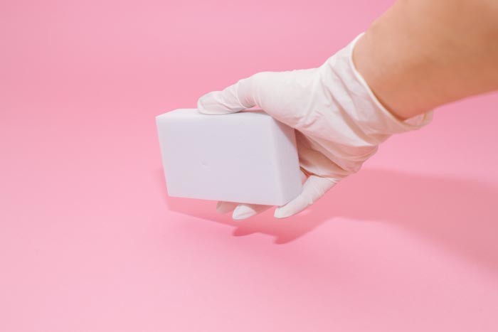 A gloved hand holding a white magic eraser against a pink background, showcasing dumb work rules.