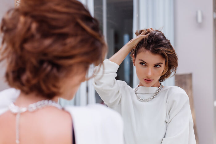 A woman with short hair looking at her reflection in a mirror, adjusting her hair, considering healthy habits.