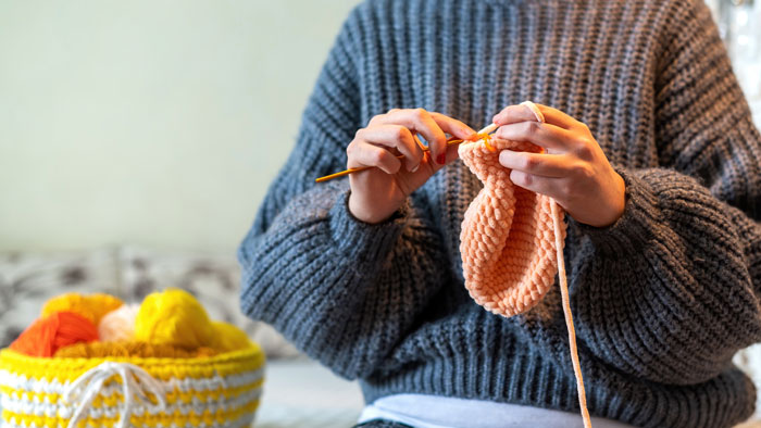 Close-up of a person in a grey sweater knitting with orange yarn, a basket of colorful yarn balls nearby, representing crisis hobbies. Close-up of a person in a grey sweater knitting with orange yarn, a basket of colorful yarn balls nearby, representing crisis hobbies.