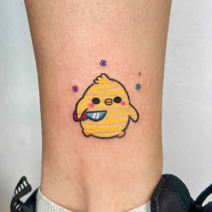 Cute yellow chick tattoo holding a knife, designed in a crayon drawing style, on an ankle.