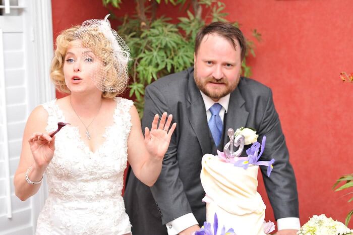 A bride gestures humorously while the groom stands beside her, next to a wedding cake, capturing funny wedding photos.