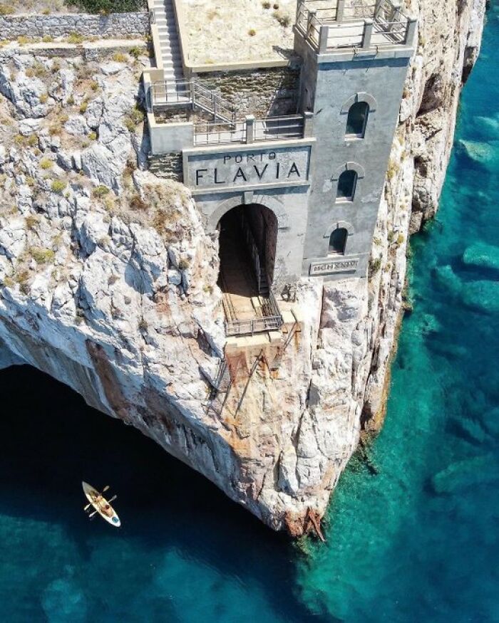 Porto Flavia, a historic building carved into a cliffside overlooking turquoise water with a small kayak below. A building with so much character.