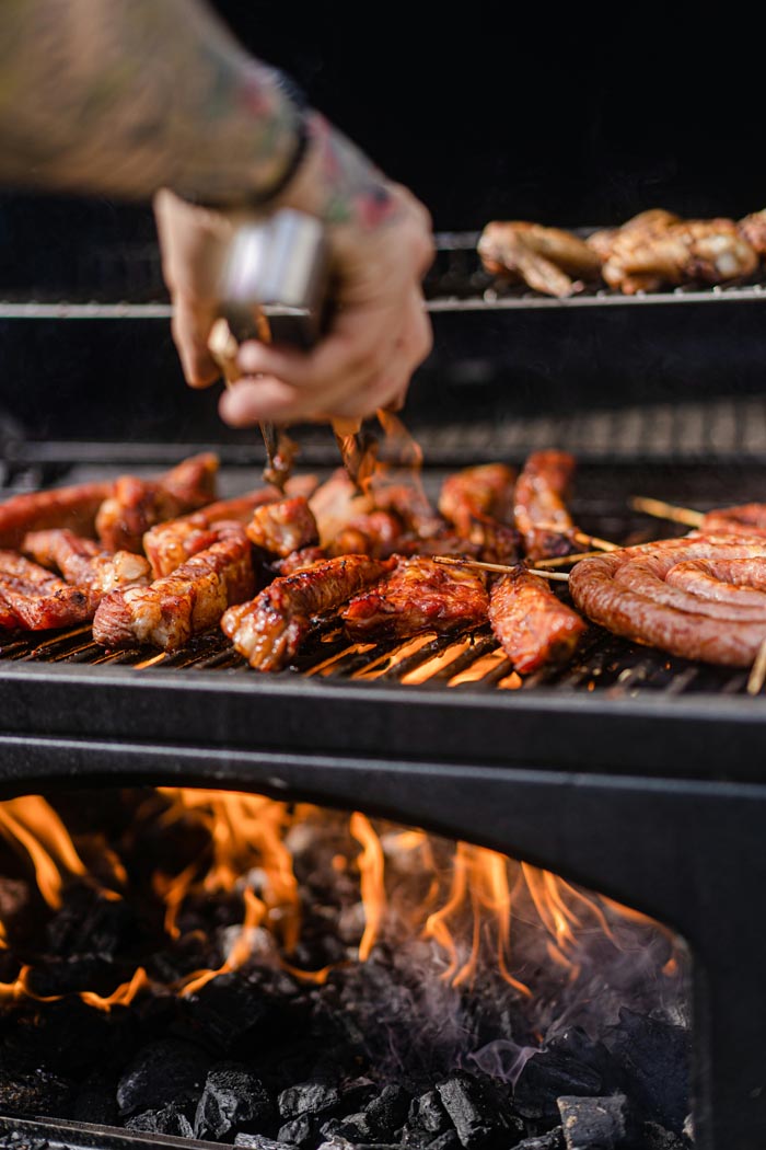 A hand using tongs to turn various meats on a hot grill with visible flames, related to dumb work rules.