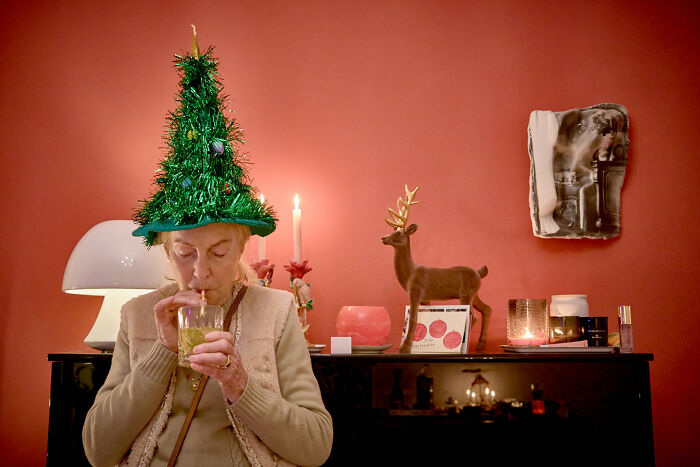 A woman wearing a Christmas tree hat sips a drink, a comical street photography scene in art.