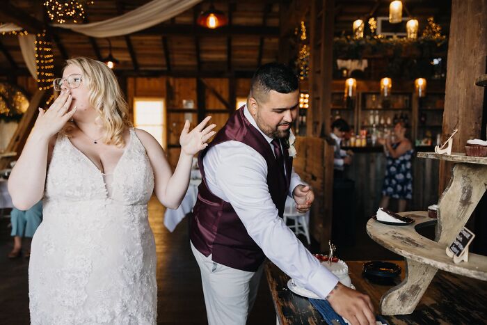 Funny wedding photo of a bride reacting to a groom cutting cake in a rustic barn.