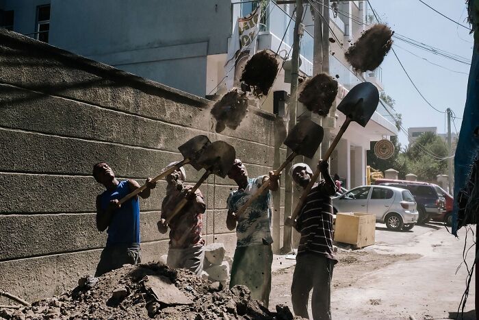 Several men in a line throwing shovelfuls of dirt into the air against an urban backdrop, capturing raw street photography.