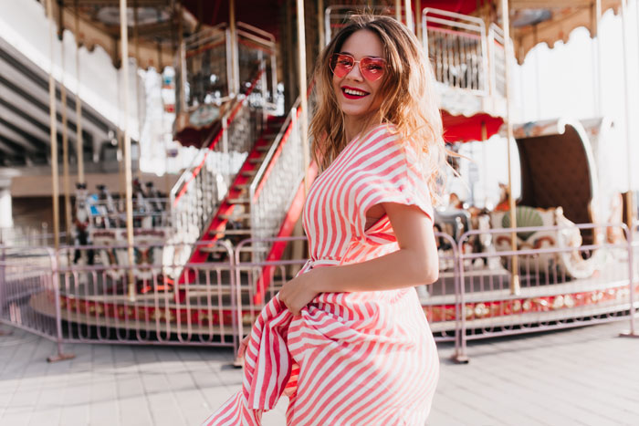 A smiling woman with wavy hair and heart-shaped sunglasses, enjoying a moment in front of a carousel, embracing millennial crisis hobbies. A smiling woman with wavy hair and heart-shaped sunglasses, enjoying a moment in front of a carousel, embracing millennial crisis hobbies.