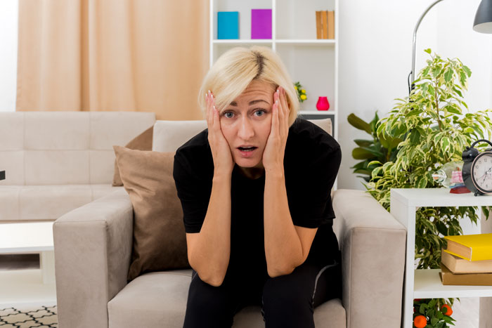 A shocked woman with blonde hair, dressed in black, sits on a sofa, expressing surprise at the expectation to house niece. A shocked woman with blonde hair, dressed in black, sits on a sofa, expressing surprise at the expectation to house niece.