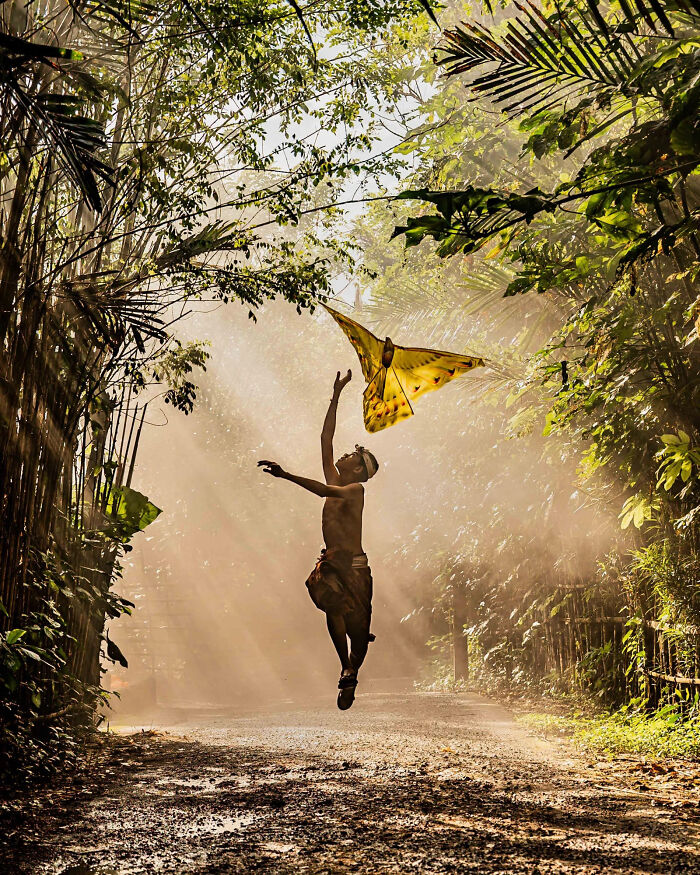 A child joyfully leaping towards a large yellow kite in a sun-drenched, lush green pathway. A winning image from the 2026 European Photography Awards.