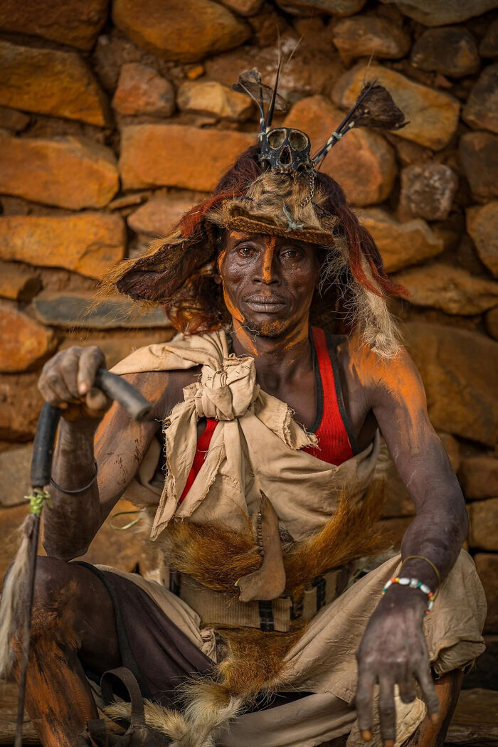 A man with intricate body paint and traditional headdress, holding a staff, posing against a stone wall, a winning image from the European Photography Awards.