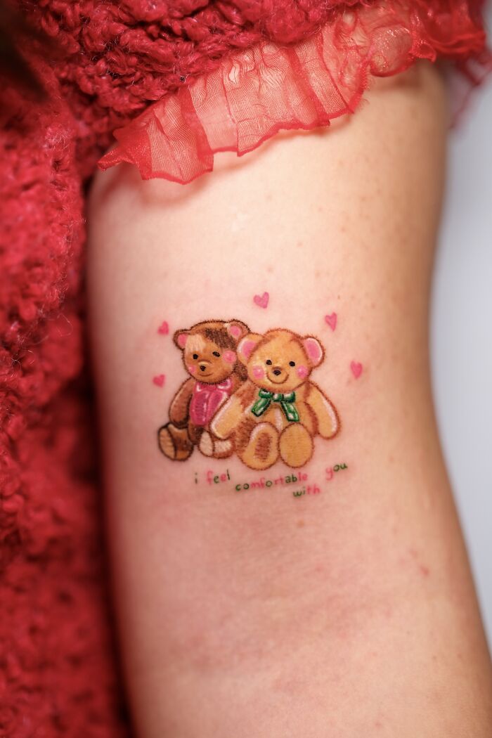 Two adorable teddy bear tattoos with hearts and a quote, resembling childhood crayon drawings, on an arm.