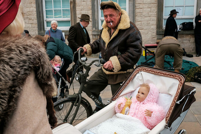 A man with a cigarette pushes a bicycle with a baby in a pram, a humorous street photography award moment.