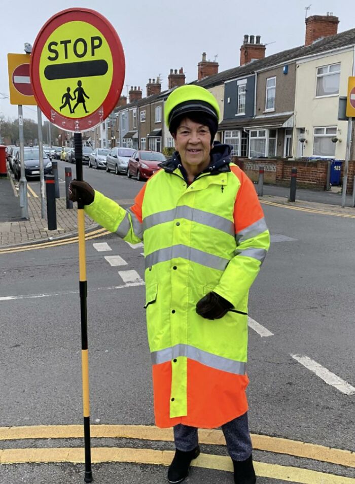 A smiling crossing guard in a high-visibility uniform holding a stop sign, embodying British humor and community spirit.