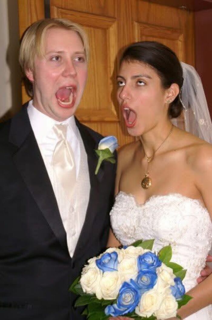 A bride and groom making exaggerated faces for a funny wedding photo, holding a bouquet of blue and white roses.