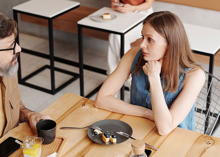 A man and a woman at a cafe table, the woman looking thoughtful as if discussing worst first dates with women.