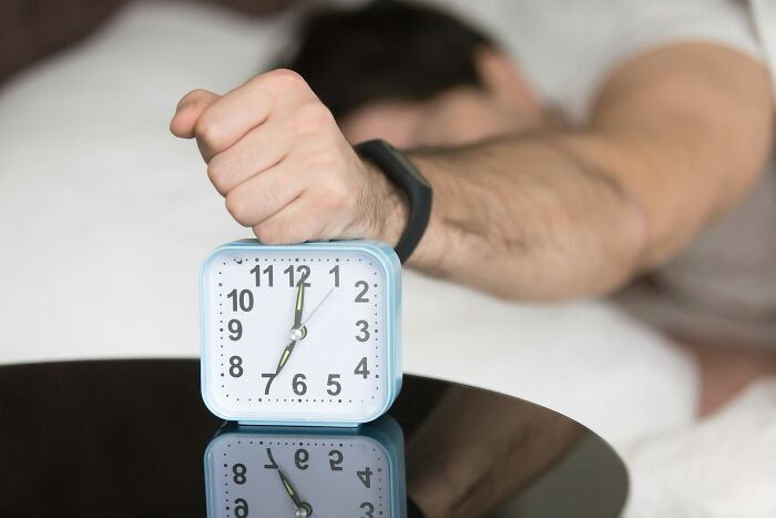 A man's hand shutting off a morning alarm clock on a table, symbolizing disrupted sleep or habits.
