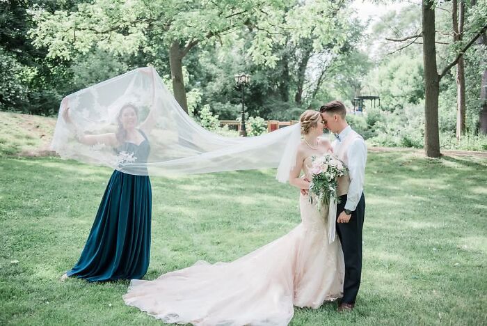 Funny wedding photo featuring a bridesmaid playfully holding the bride's veil over the kissing couple, an iconic moment.