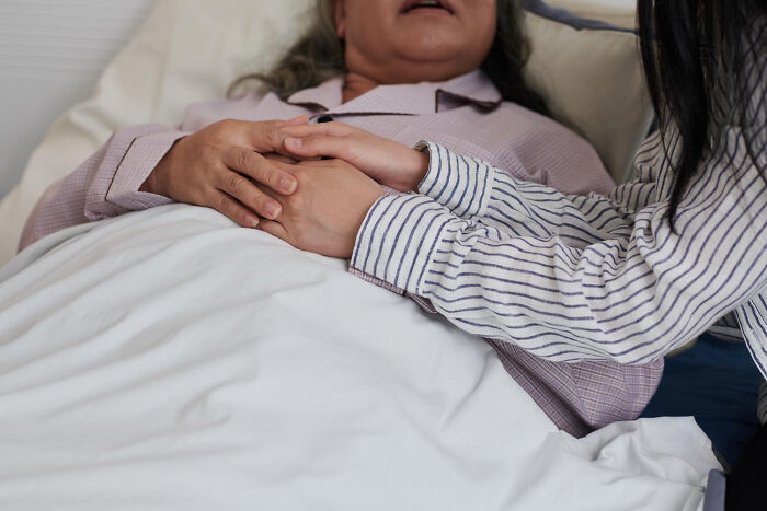 Two people holding hands, one in a hospital bed, suggesting a shared secret or confession.