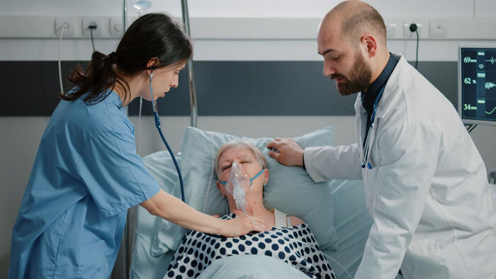 A female nurse and male doctor tend to an elderly female patient in a hospital bed, who is wearing an oxygen mask, in the medical field.