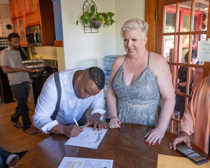 Funny wedding photo of a groom signing documents while a woman in a grey dress watches, an iconic moment.