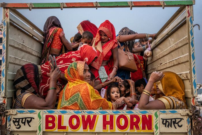 Many women and children are crowded in the back of a truck, featuring a Blow Horn sign, a street photo capturing life's coincidences.